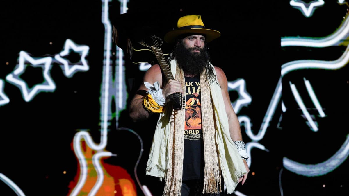 Elijah in a hat,scarf, and cut off tee with a guitar slung over his shoulder, stands on a TNA Wrestling stage