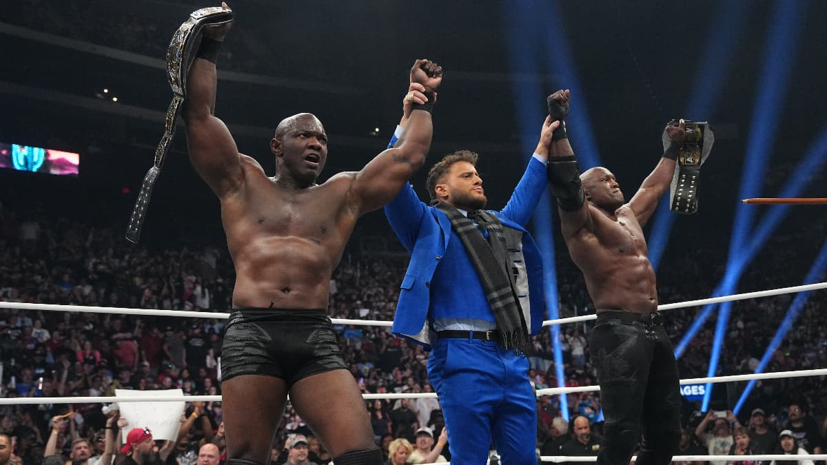 MJF in a blue suit raises the hands of Shelton Benjamin and Bobby Lashley in a wrestling ring, the latter two men holding wrestling championship belts