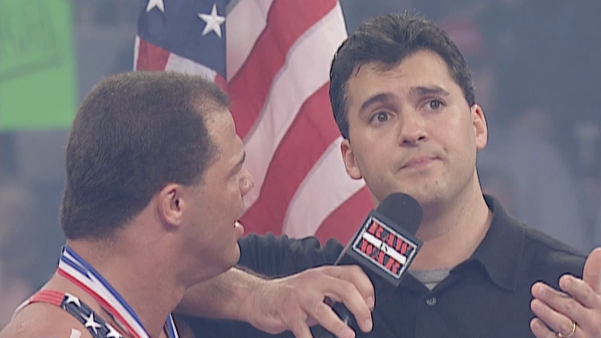 Kurt Angle looking on annoyed at Shane McMahon, who is resting on his shoulder holding a WWF Raw is War microphone