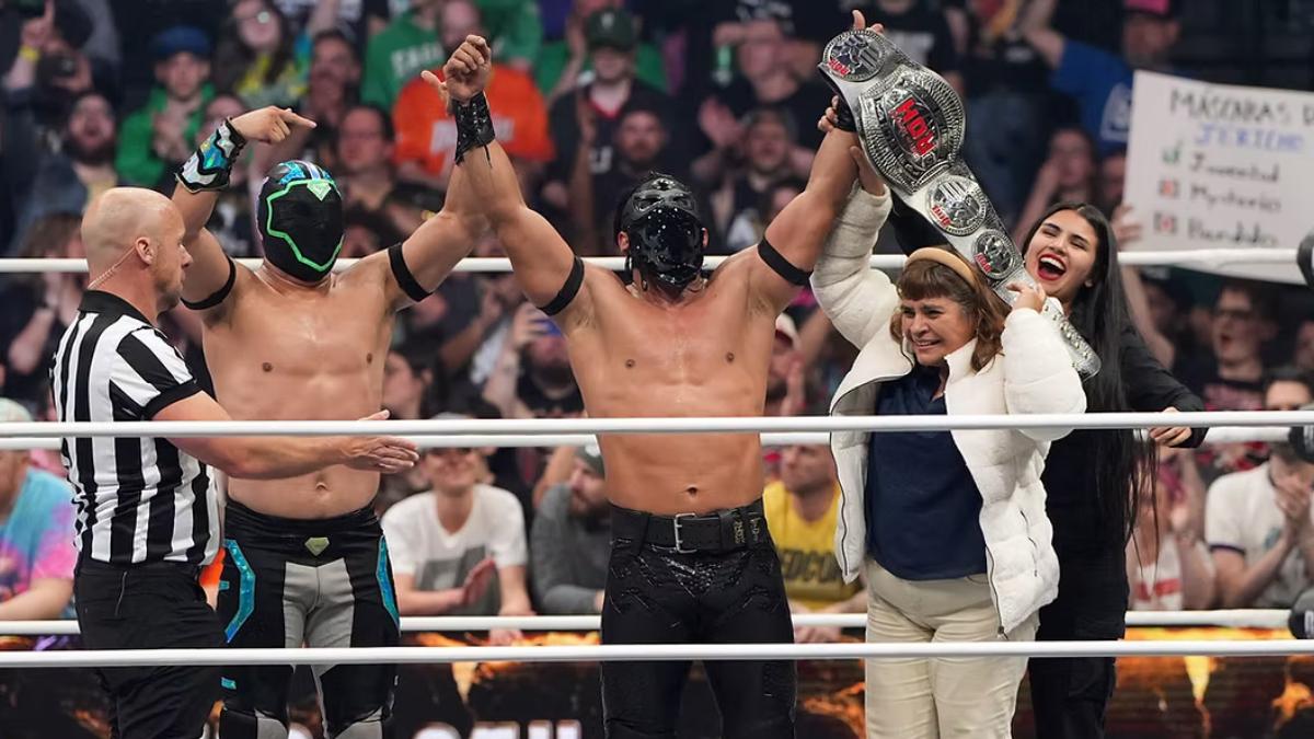 Bandido holds up the ROH World Championship in a wrestling ring, flanked by brother Gravity to his right, and his mother and sister to his left
