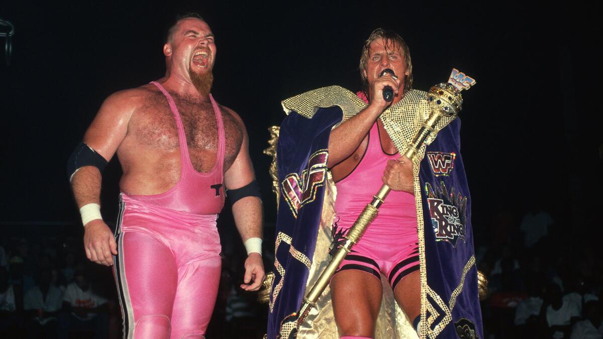 Owen Hart cutting a promo while wearing a cape and holding a sceptre alongside Jim Neidhart, who is in all-pink attire