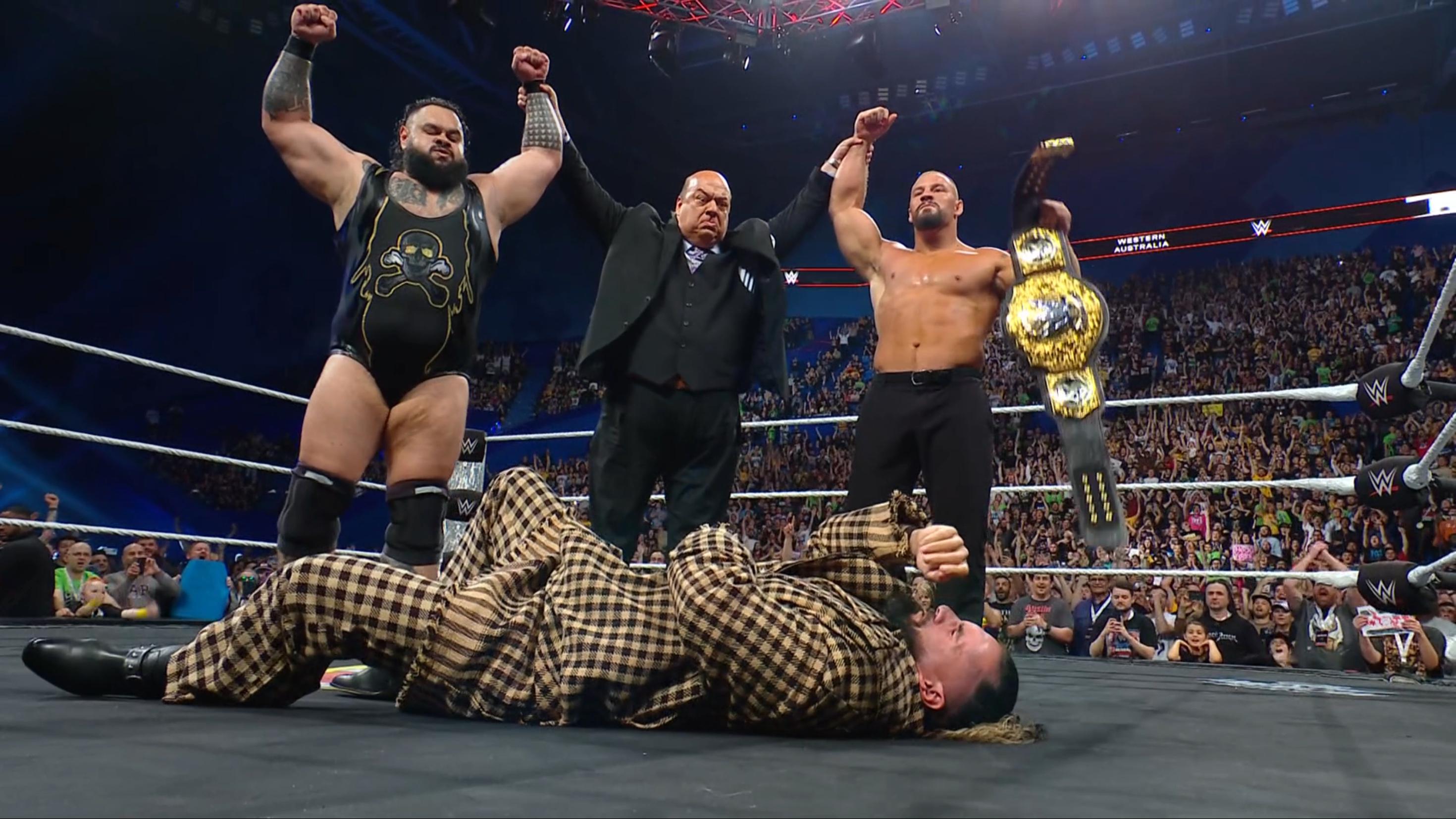Seth Rollins lying down as Paul Heyman raises Bronson Reed and Bron Breakker's arms, with Breakker also holding the WWE World Heavyweight Title