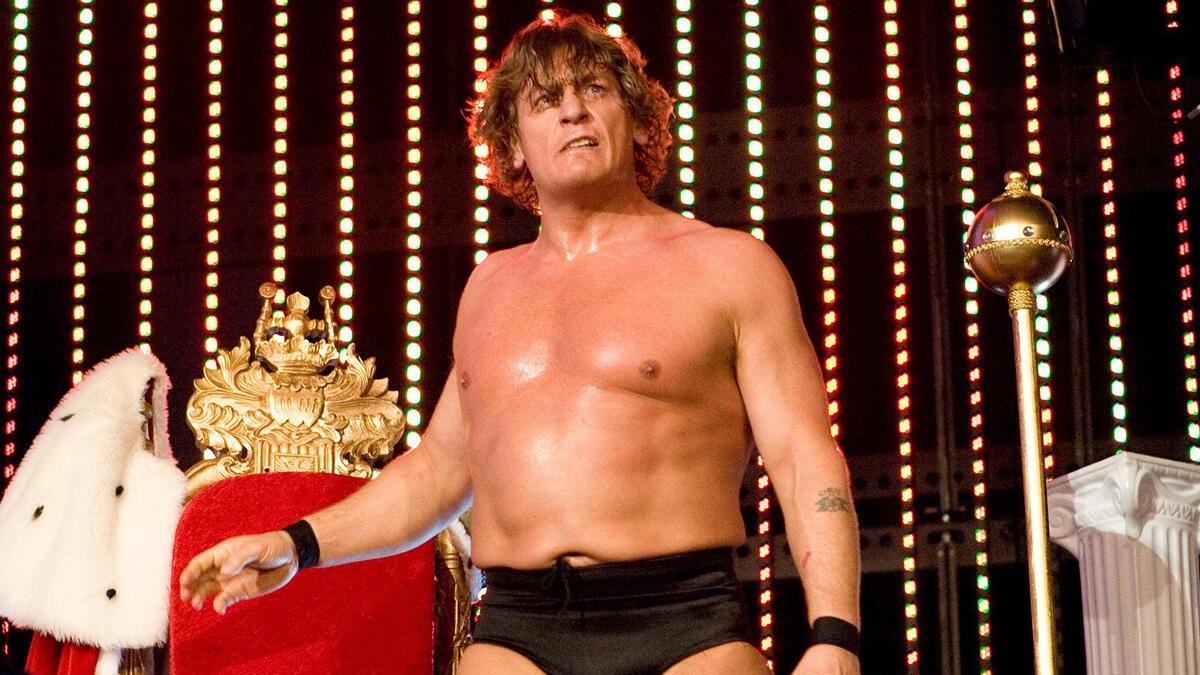 William Regal standing next to a throne in 2008 while wearing black trunks and with a mop of hair